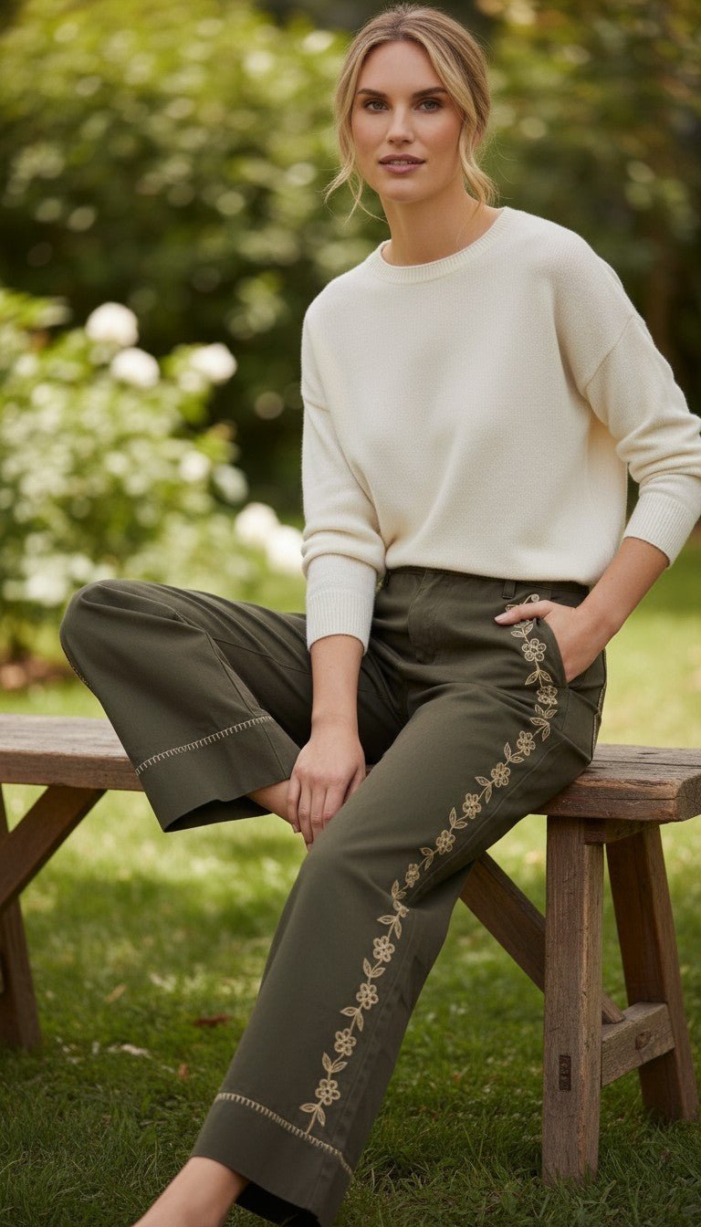 Model wearing graced olive green cotton wide-leg pants with floral embroidery and contrast stitching on a wooden bench