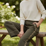 Model wearing graced olive green cotton wide-leg pants with floral embroidery and contrast stitching on a wooden bench