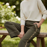 Model wearing graced olive green cotton wide-leg pants with floral embroidery and contrast stitching on a wooden bench