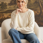 A woman wearing a graced ivory Eloise crew neck sweater with dropped shoulders, sitting in a chair against a landscape painting.