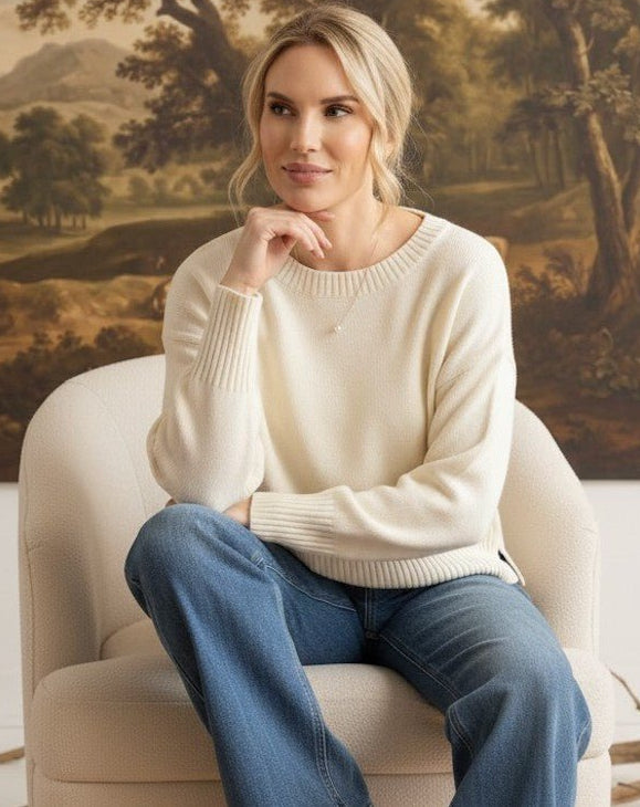 A woman wearing a graced ivory Eloise crew neck sweater with dropped shoulders, sitting in a chair against a landscape painting.