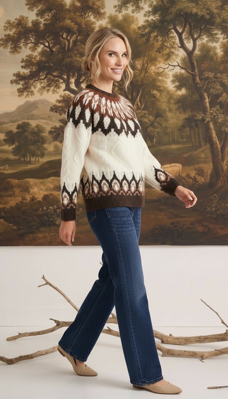 A woman wearing a graced Fair Isle cable knit sweater in cream and brown with blue jeans against a landscape backdrop.