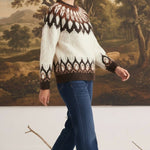 A woman wearing a graced Fair Isle cable knit sweater in cream and brown with blue jeans against a landscape backdrop.