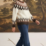 A woman wearing a graced Fair Isle cable knit sweater in cream and brown with blue jeans against a landscape backdrop.