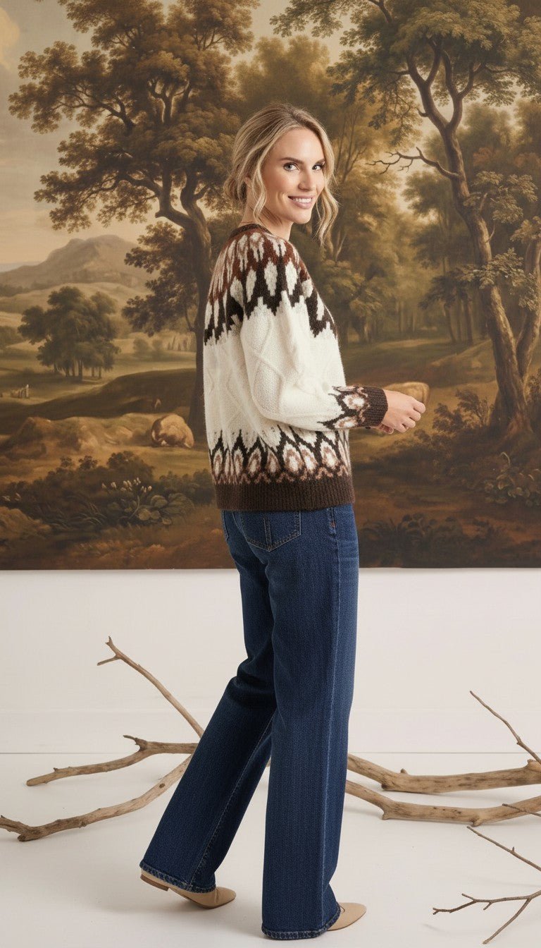 A woman wearing a graced Fair Isle cable knit sweater in cream and brown with blue jeans, standing in a studio.