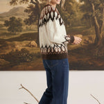 A woman wearing a graced Fair Isle cable knit sweater in cream and brown with blue jeans, standing in a studio.