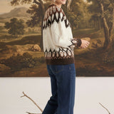 A woman wearing a graced Fair Isle cable knit sweater in cream and brown with blue jeans, standing in a studio.