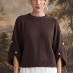 A woman stands indoors wearing a dark brown knit sweater with wide draped sleeves and decorative pearl-like buttons on the cuffs, paired with white shorts. A scenic landscape tapestry is visible behind her.