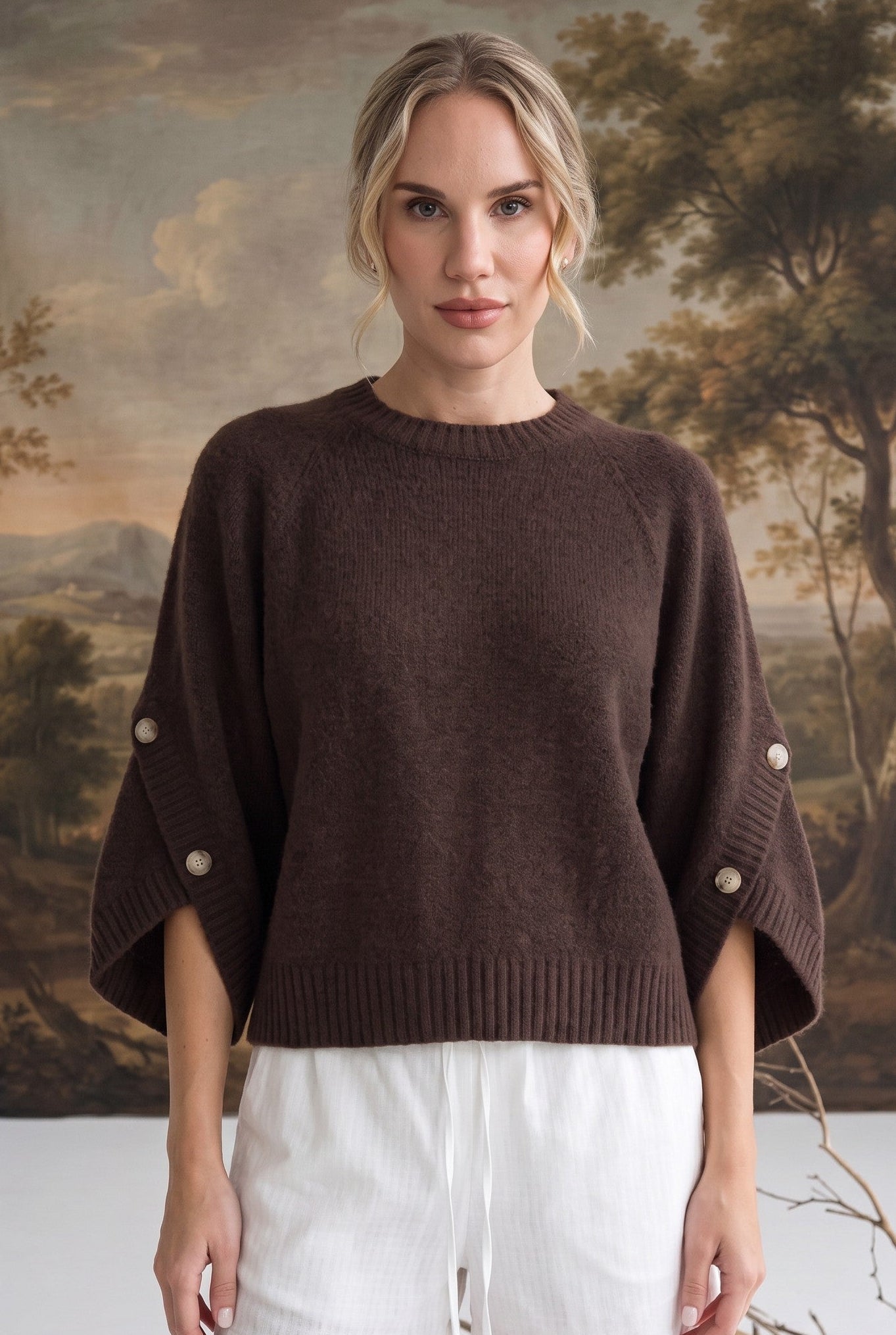 A woman stands indoors wearing a dark brown knit sweater with wide draped sleeves and decorative pearl-like buttons on the cuffs, paired with white shorts. A scenic landscape tapestry is visible behind her.