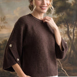 A woman wearing a brown knit sweater with wide three-quarter sleeves and decorative buttons, paired with white pants, standing in front of a large landscape tapestry.