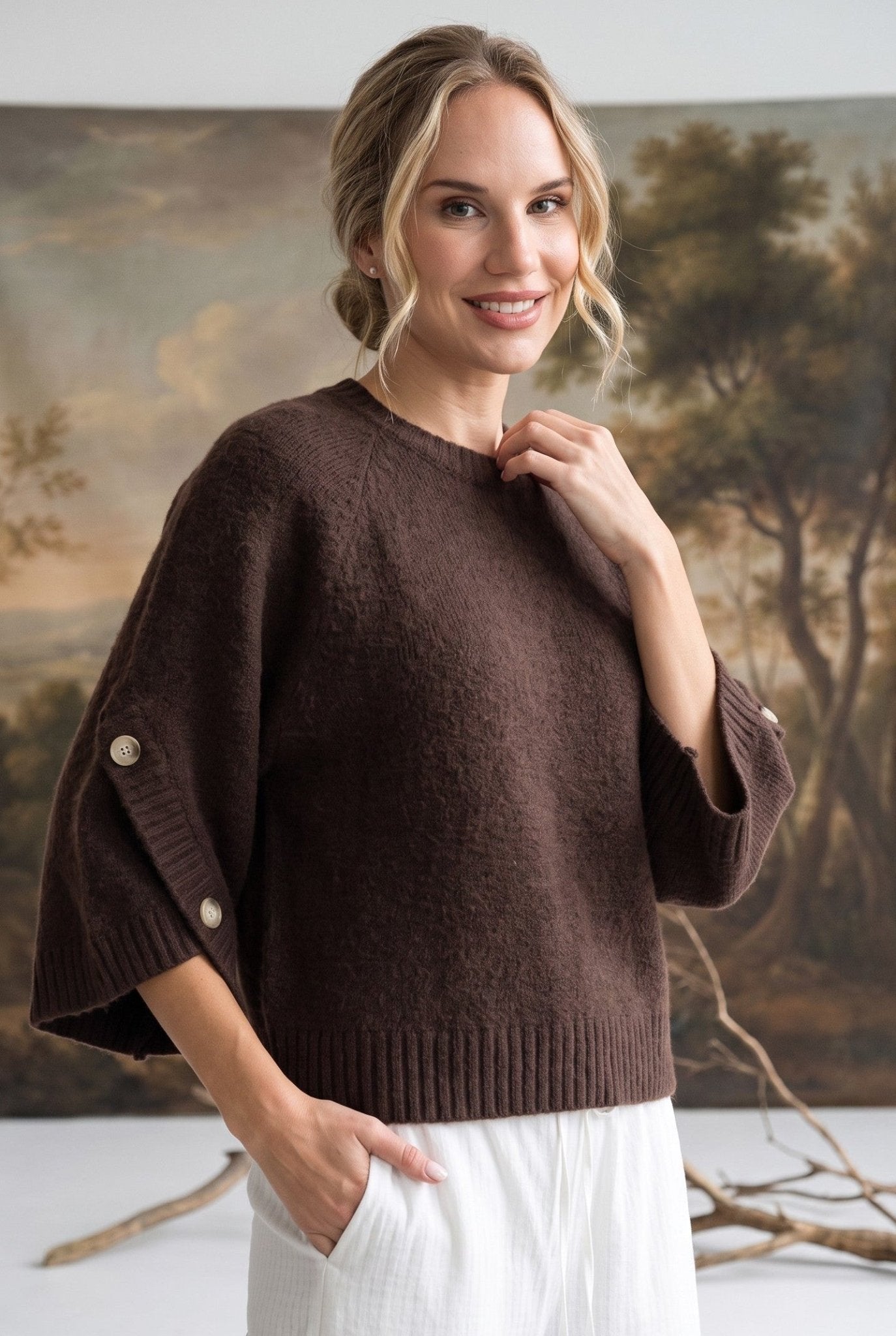 A woman wearing a brown knit sweater with wide three-quarter sleeves and decorative buttons, paired with white pants, standing in front of a large landscape tapestry.