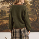 graced The Lola Pocket Detail Cardigan in olive green with brown trim, worn by a woman with a plaid skirt