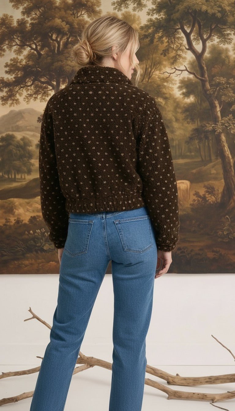 Back view of a woman wearing a brown patterned fleece jacket from graced with blue jeans in a studio setting.