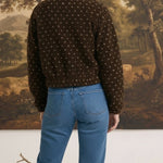 Back view of a woman wearing a brown patterned fleece jacket from graced with blue jeans in a studio setting.