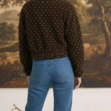 Back view of a woman wearing a brown patterned fleece jacket from graced with blue jeans in a studio setting.