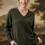 A woman modeling a dark olive green V-neck sweater and black pants, standing in front of a large classical landscape painting.