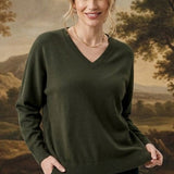 A woman modeling a dark olive green V-neck sweater and black pants, standing in front of a large classical landscape painting.