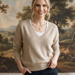 A woman stands indoors wearing a beige v-neck knit sweater over a white top and dark navy pants, with a large landscape painting hanging on the wall behind her.