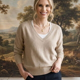 A woman stands indoors wearing a beige v-neck knit sweater over a white top and dark navy pants, with a large landscape painting hanging on the wall behind her.