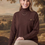 graced Remy brown turtleneck sweater with a single chest pocket, worn by a smiling woman in a rustic landscape setting