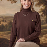 graced Remy brown turtleneck sweater with a single chest pocket, worn by a smiling woman in a rustic landscape setting