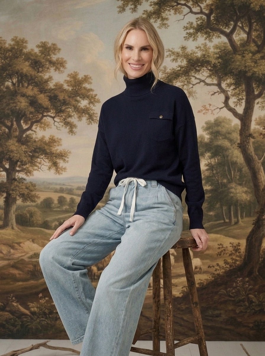 graced Remy turtleneck sweater in navy with a chest pocket, worn by a woman sitting on a wooden stool