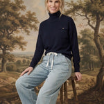 graced Remy turtleneck sweater in navy with a chest pocket, worn by a woman sitting on a wooden stool