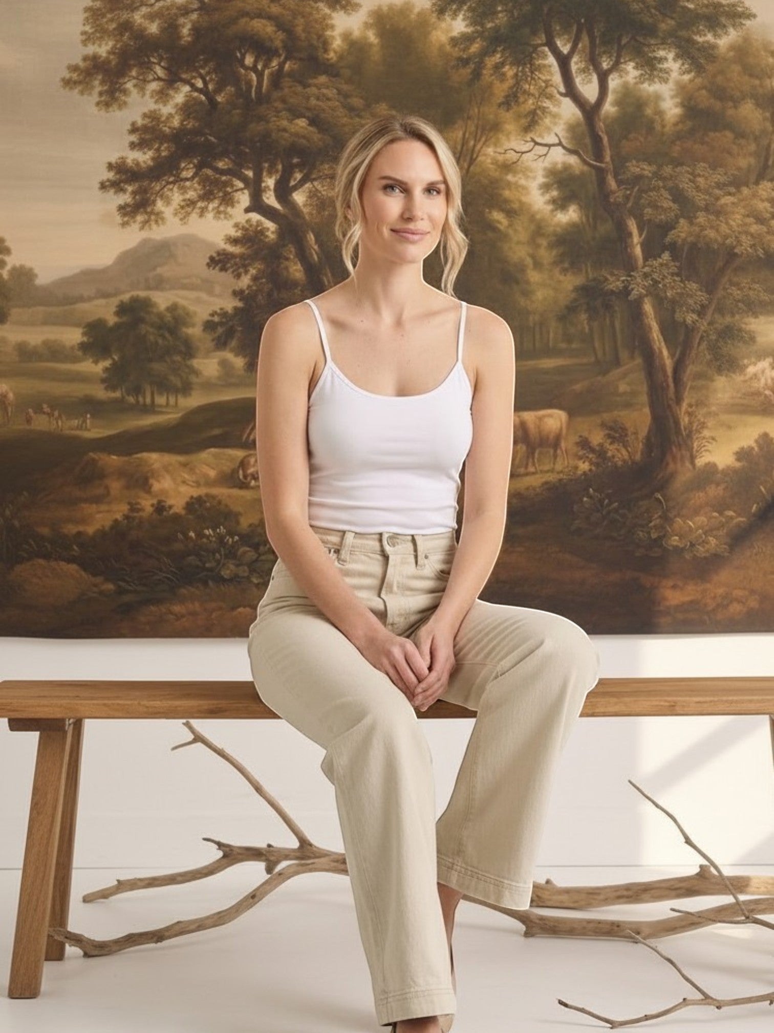 graced model wearing a white fitted spaghetti strap cami with beige jeans, sitting on a wooden bench
