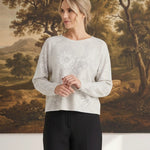 A woman wearing a graced Susie diamond pointelle floral sweater in light grey with delicate knit patterns.