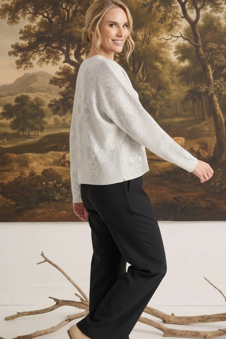 A woman wearing a graced Susie diamond pointelle floral sweater in white, paired with black trousers, side view.