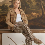 Model wearing graced Tina plaid checker pants in brown and white with a denim jacket, sitting on a wooden bench.